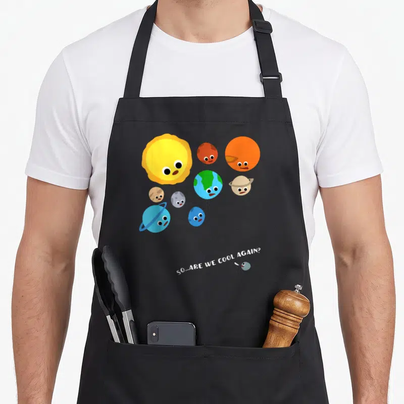 Versus planets apron - Cartoon planets cluster around sun with diagonal text asking about Pluto below — shown on a black cotton canvas apron against a white background