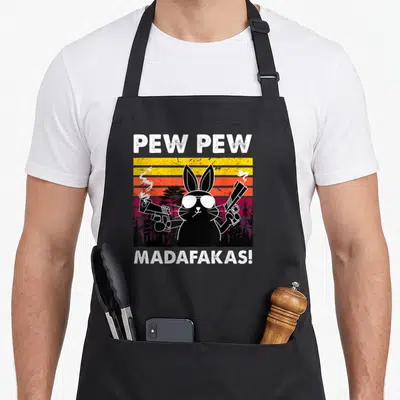 Pew bunny apron - Black rabbit with sunglasses and guns on a striped yellow orange purple sunset background — shown on a black cotton canvas apron against a white background