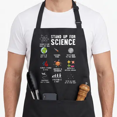 Science change apron - Stand Up For Science text with icons for Earth, vaccines, moon, climate, math, evolution — shown on a black cotton canvas apron against a white background