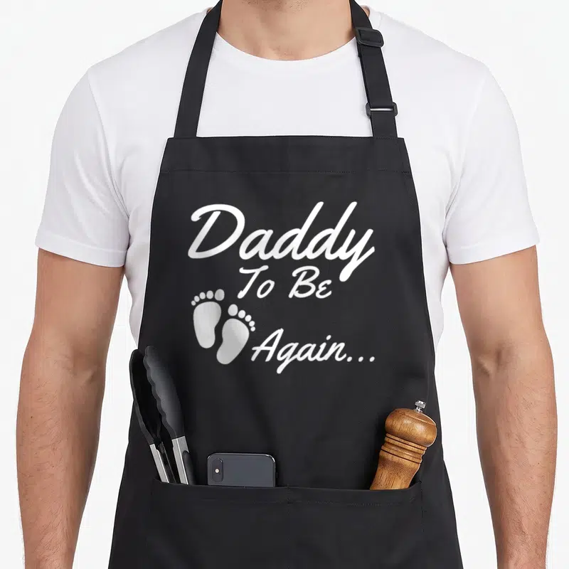 Funny soon daddy apron - White 'Daddy To Be Again...' text with two baby footprints on left — shown on a black cotton canvas apron against a white background