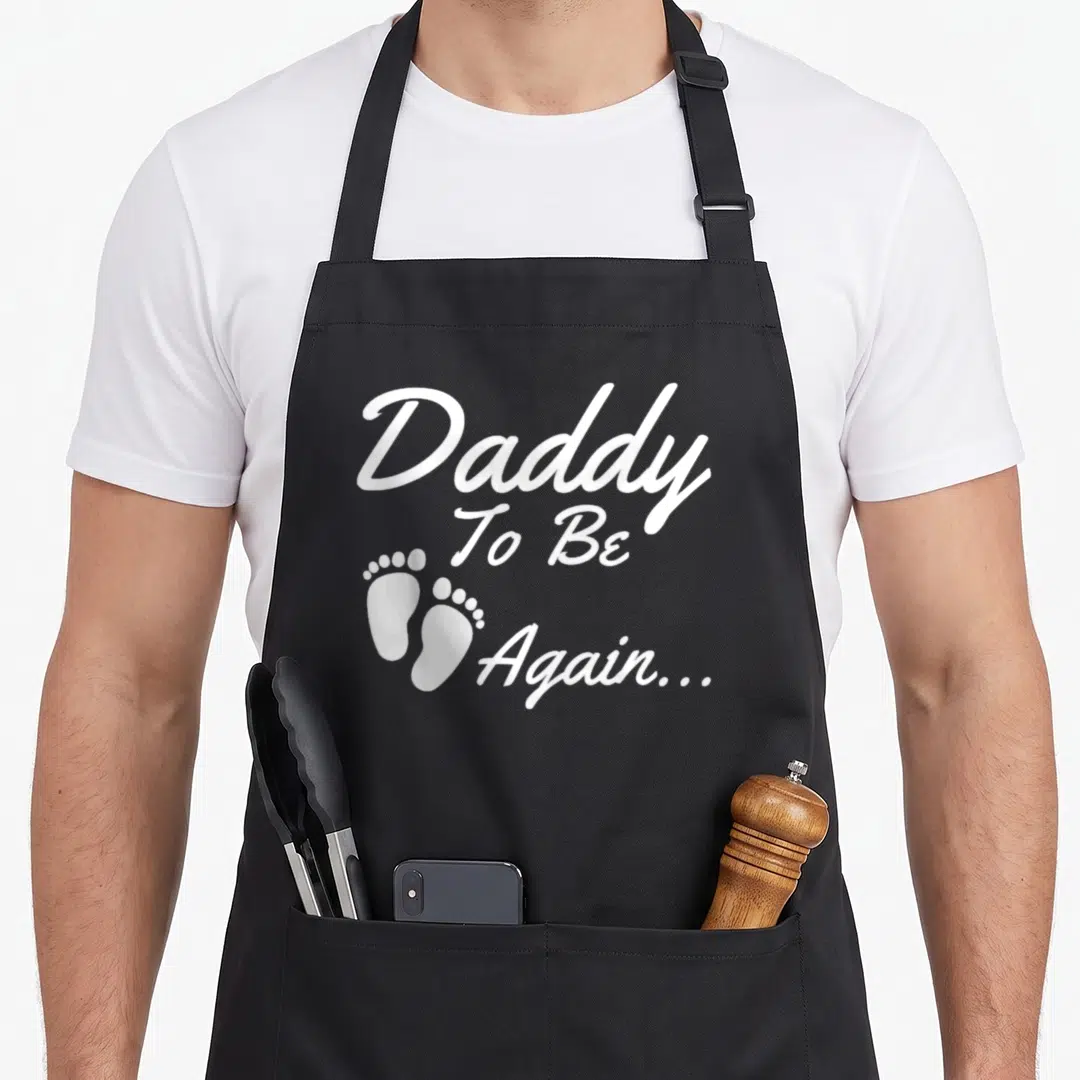 Funny soon daddy apron - White 'Daddy To Be Again...' text with two baby footprints on left — shown on a black cotton canvas apron against a white background