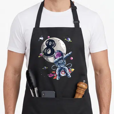 Space 8th birthday apron - Cartoon astronaut on rocket in front of moon with black number 8 and planets — shown on a black cotton canvas apron against a white background
