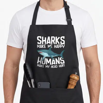 Shark studying life apron - Blue cartoon shark centered between white text reading sharks make me happy and humans make — shown on a black cotton canvas apron against a white background