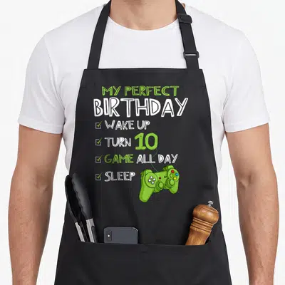 Birthday gaming aprons - Lime green controller connected by dotted line to birthday checklist text on black fabric — shown on a black cotton canvas apron against a white background