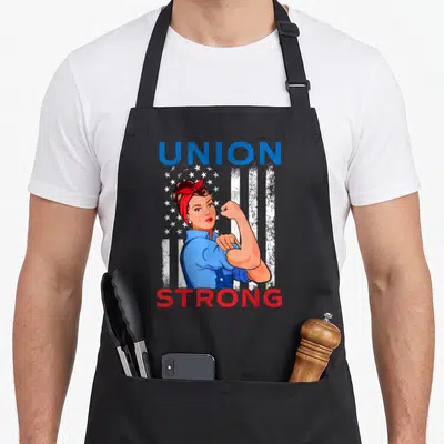 Strong labor union aprons - Rosie the Riveter flexing arm with UNION STRONG text over stylized flag in blue red white — shown on a black cotton canvas apron against a white background