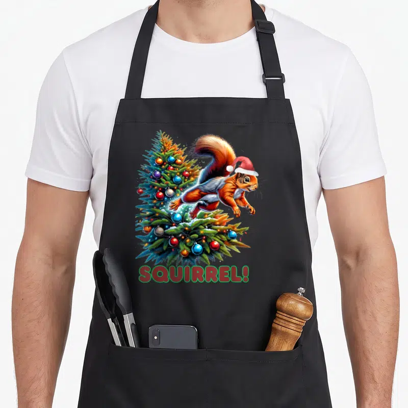 Bean christmas tree apron - Cartoon squirrel in red santa hat jumping past green tree with blue red and orange — shown on a black cotton canvas apron against a white background