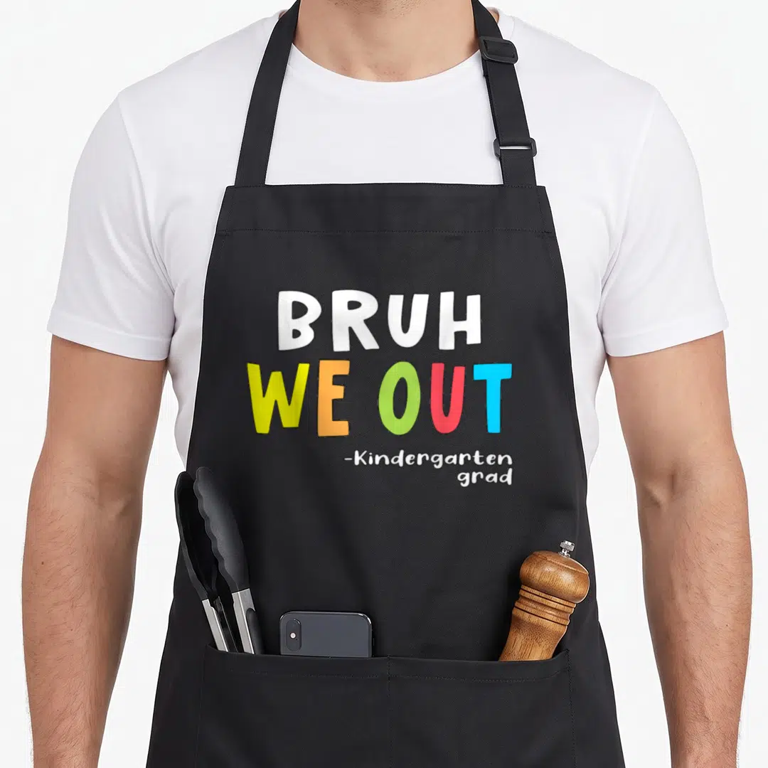 Goodbye kindergarten hello apron - BRUH WE OUT text in white yellow orange green red blue stacked vertically — shown on a black cotton canvas apron against a white background