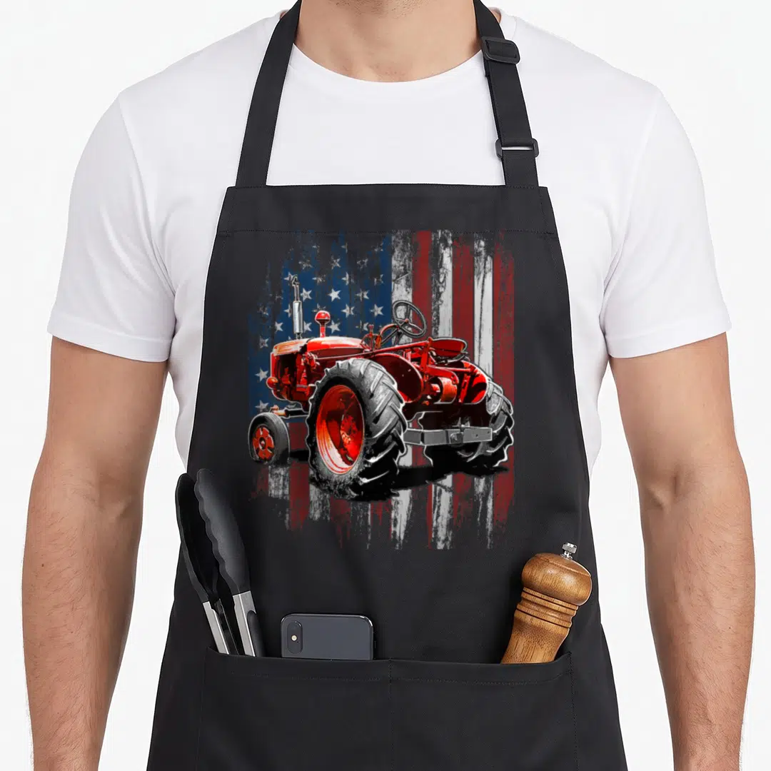 Farmer american flag apron - Red tractor illustration in front of a weathered American flag pattern — shown on a black cotton canvas apron against a white background