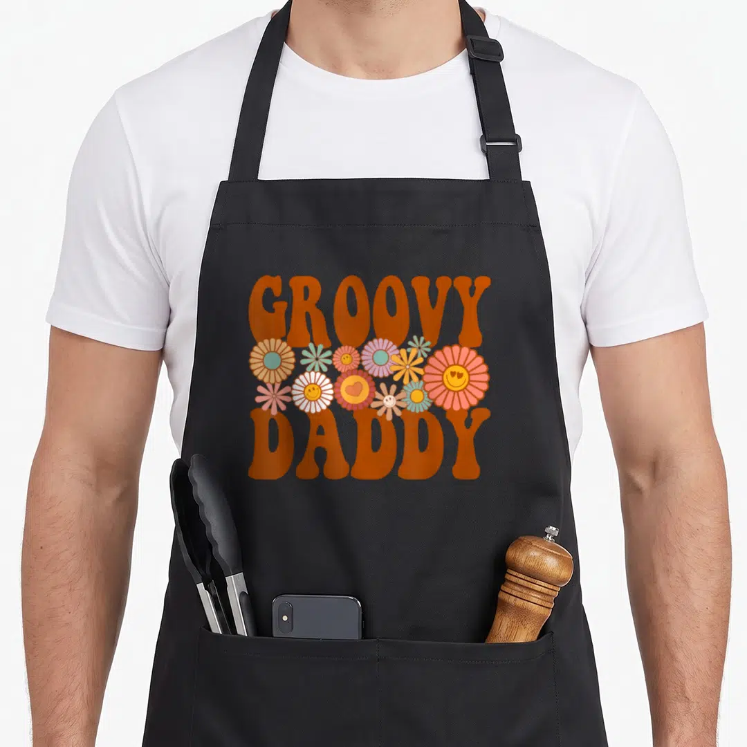 Family daddy apron - Retro GROOVY DADDY text with smiley face flowers in orange, pink, and yellow — shown on a black cotton canvas apron against a white background