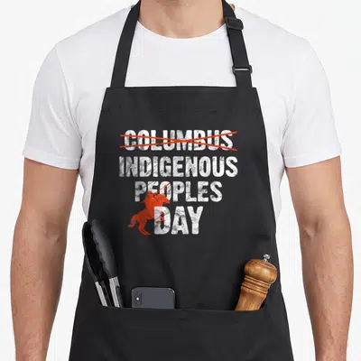 Columbus indigenous apron - Stacked white text with Columbus crossed out in red and a red horse silhouette overlay — shown on a black cotton canvas apron against a white background