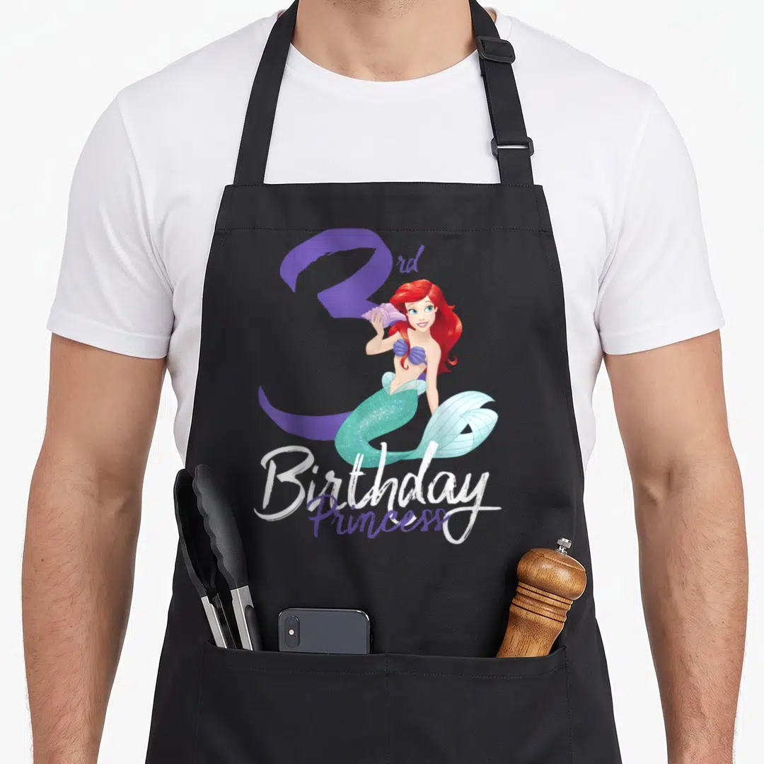 3rd princess apron - Cartoon mermaid with red hair and teal tail in front of purple number three with Birthday Princess — shown on a black cotton canvas apron against a white background