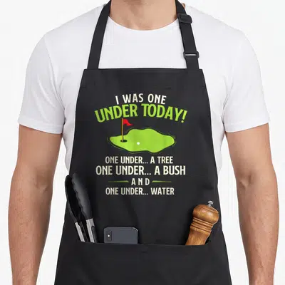 Golfing golf apron - I Was One Under Today text over lime green golf green with red flag and white ball — shown on a black cotton canvas apron against a white background