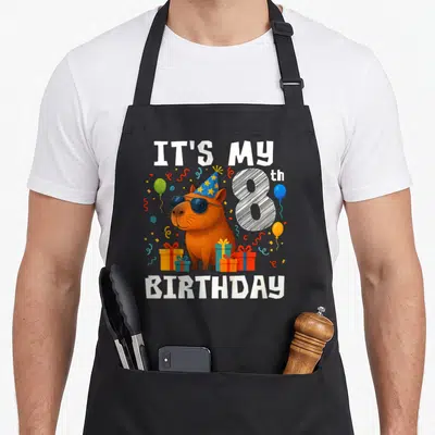 Cute 8th birthday apron - Brown capybara in sunglasses and star hat with gifts, balloons, and IT'S MY 8th BIRTHDAY text — shown on a black cotton canvas apron against a white background