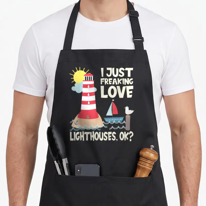 Lighthouse freaking love apron - Cartoon red white lighthouse on tan island with boat seagull and text I just freaking — shown on a black cotton canvas apron against a white background