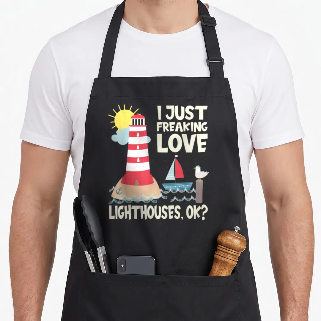 Lighthouse freaking love apron - Cartoon red white lighthouse on tan island with boat seagull and text I just freaking — shown on a black cotton canvas apron against a white background