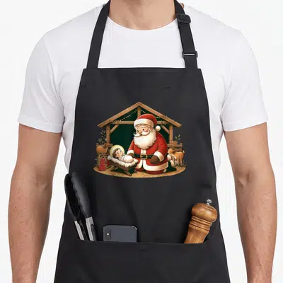 Kneeling santa claus apron - Cartoon Santa kneeling beside baby Jesus in a stable with two goats in red and brown — shown on a black cotton canvas apron against a white background
