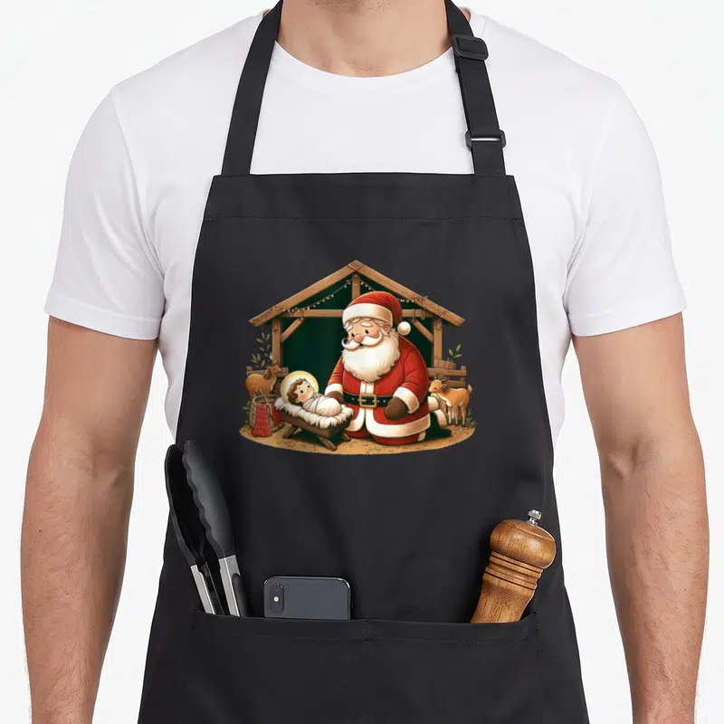 Kneeling santa claus apron - Cartoon Santa kneeling beside baby Jesus in a stable with two goats in red and brown — shown on a black cotton canvas apron against a white background