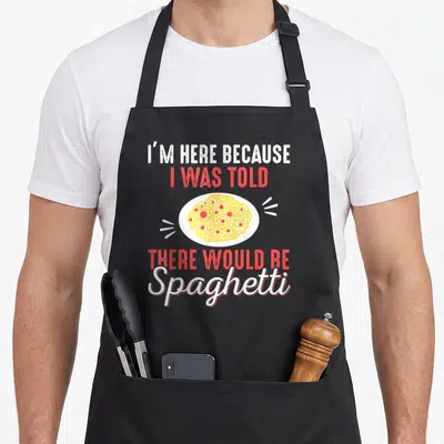 Funny spaghetti pasta apron - I'm Here Because There Would Be Spaghetti text surrounding cartoon noodle plate — shown on a black cotton canvas apron against a white background