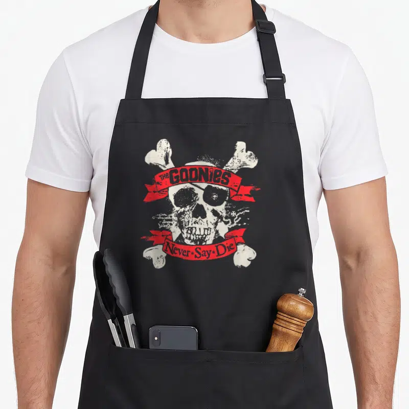 Crossbone skull apron - Distressed white skull with eyepatch over crossed bones. Red ribbons read The Goonies Never Say — shown on a black cotton canvas apron against a white background