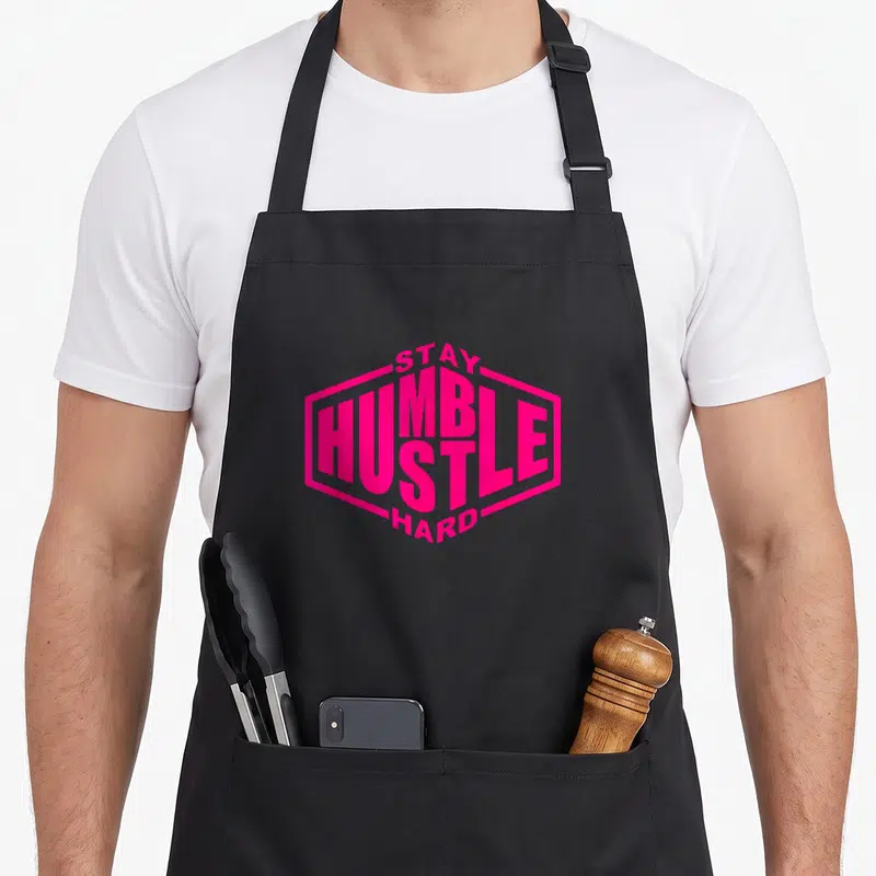 Stay urban apron - Pink text STAY HUMBLE HUSTLE HARD in hexagon border — shown on a black cotton canvas apron against a white background