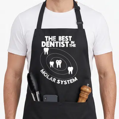 System dentist apron - White text THE BEST DENTIST IN THE MOLAR SYSTEM with four teeth orbiting a smiling center tooth — shown on a black cotton canvas apron against a white background