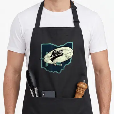 Akron distressed apron - Cream blimp with Akron text over dark blue Ohio map outline with teal border — shown on a black cotton canvas apron against a white background