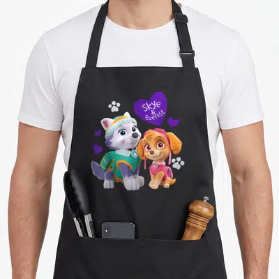 Everest isle of skye apron - Two cartoon dogs sit near a purple heart with Skye & Everest text and glittery paws — shown on a black cotton canvas apron against a white background