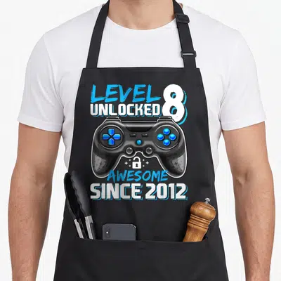 Level unlocked 8th video game apron - Game controller with blue buttons, small lock, plus signs, and stacked text — shown on a black cotton canvas apron against a white background