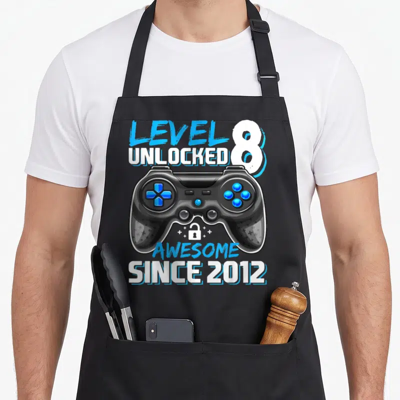 Level unlocked 8th video game apron - Game controller with blue buttons, small lock, plus signs, and stacked text — shown on a black cotton canvas apron against a white background