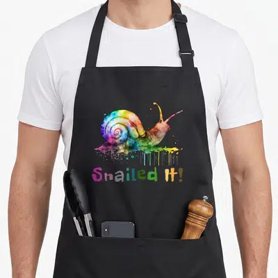 Snail rainbow apron - Watercolor rainbow snail with paint drips and Snailed It text — shown on a black cotton canvas apron against a white background