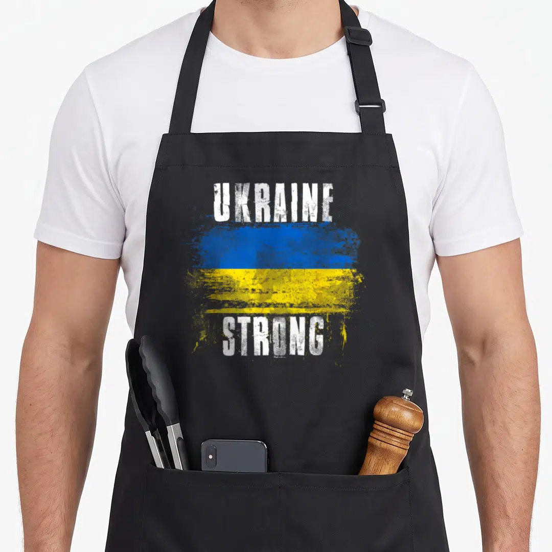 Strong ukrainian aprons - Distressed blue yellow flag with white UKRAINE STRONG text on black fabric — shown on a black cotton canvas apron against a white background