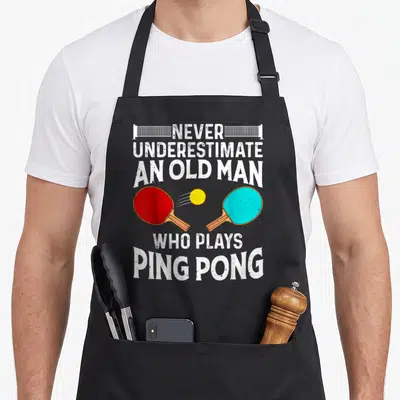 Grandpa table apron - Distressed white text above and below red and blue paddles hitting a yellow ping pong ball — shown on a black cotton canvas apron against a white background