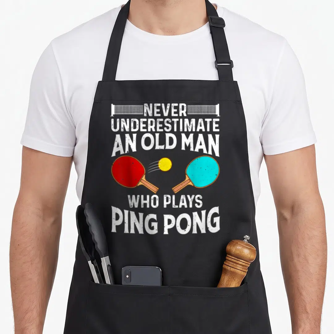 Grandpa table apron - Distressed white text above and below red and blue paddles hitting a yellow ping pong ball — shown on a black cotton canvas apron against a white background