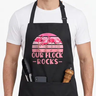 Our flock family aprons - Four pink flamingos standing before a striped sun with OUR FLOCK -ROCKS- text below — shown on a black cotton canvas apron against a white background