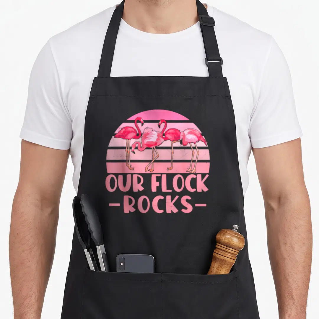 Our flock family aprons - Four pink flamingos standing before a striped sun with OUR FLOCK -ROCKS- text below — shown on a black cotton canvas apron against a white background