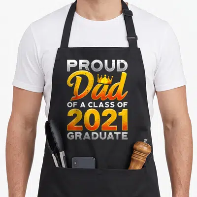 Class school dad apron - Stacked text PROUD Dad OF A CLASS OF 2021 GRADUATE with crown and yellow orange white colors — shown on a black cotton canvas apron against a white background
