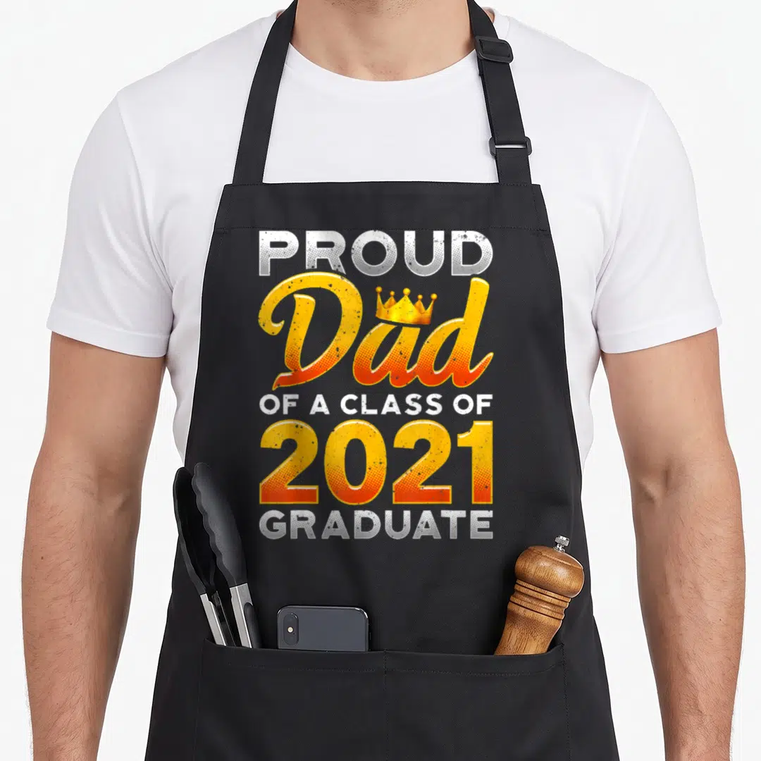 Class school dad apron - Stacked text PROUD Dad OF A CLASS OF 2021 GRADUATE with crown and yellow orange white colors — shown on a black cotton canvas apron against a white background