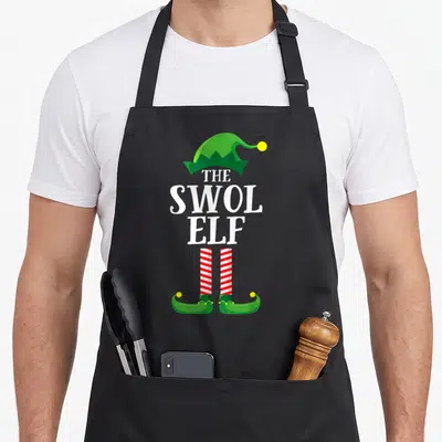 Swol christmas elf apron - Stacked white text THE SWOL ELF with green hat and striped legs — shown on a black cotton canvas apron against a white background