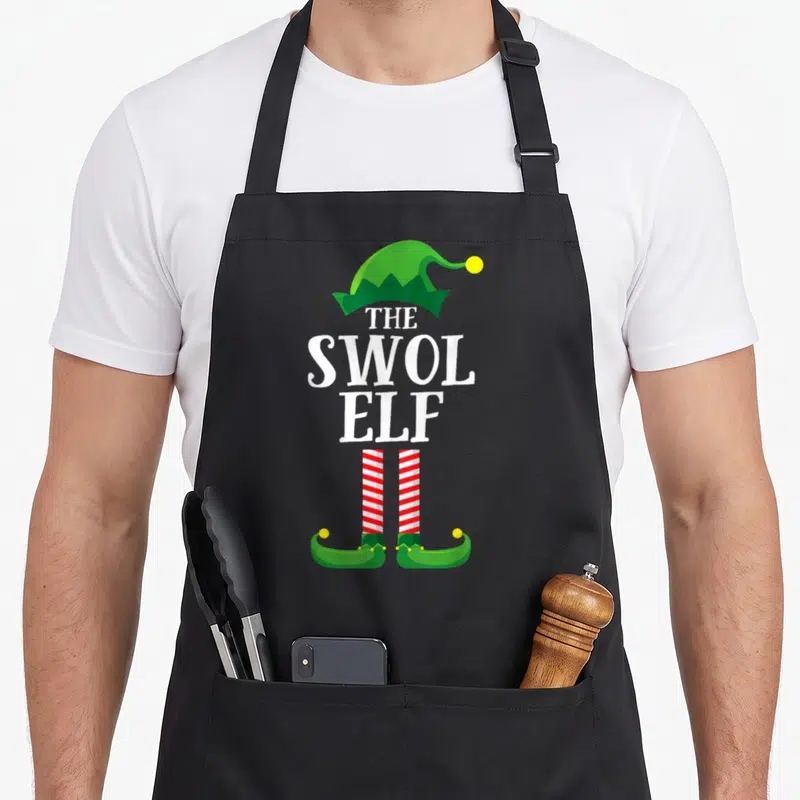 Swol christmas elf apron - Stacked white text THE SWOL ELF with green hat and striped legs — shown on a black cotton canvas apron against a white background