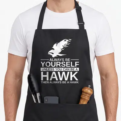 Alway bird apron - White hawk silhouette above stacked text reading Always Be Yourself Unless You Can Be A Hawk Then — shown on a black cotton canvas apron against a white background