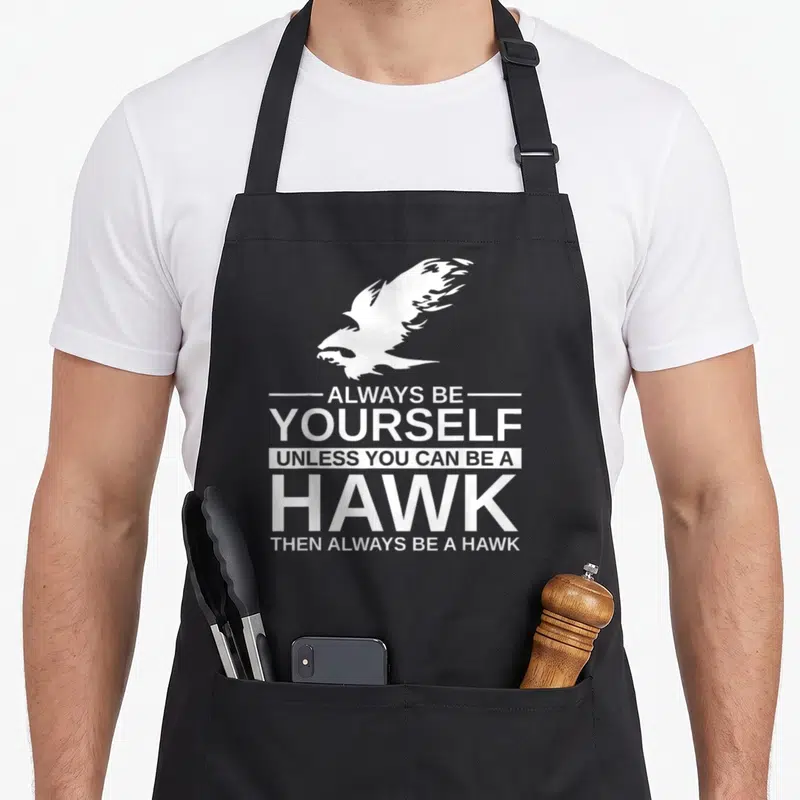 Alway bird apron - White hawk silhouette above stacked text reading Always Be Yourself Unless You Can Be A Hawk Then — shown on a black cotton canvas apron against a white background