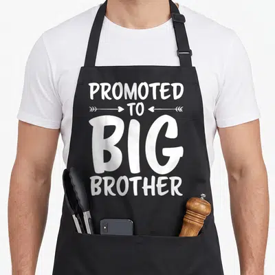 Funny brother promoted big apron - White PROMOTED TO BIG BROTHER text with two inward arrows — shown on a black cotton canvas apron against a white background