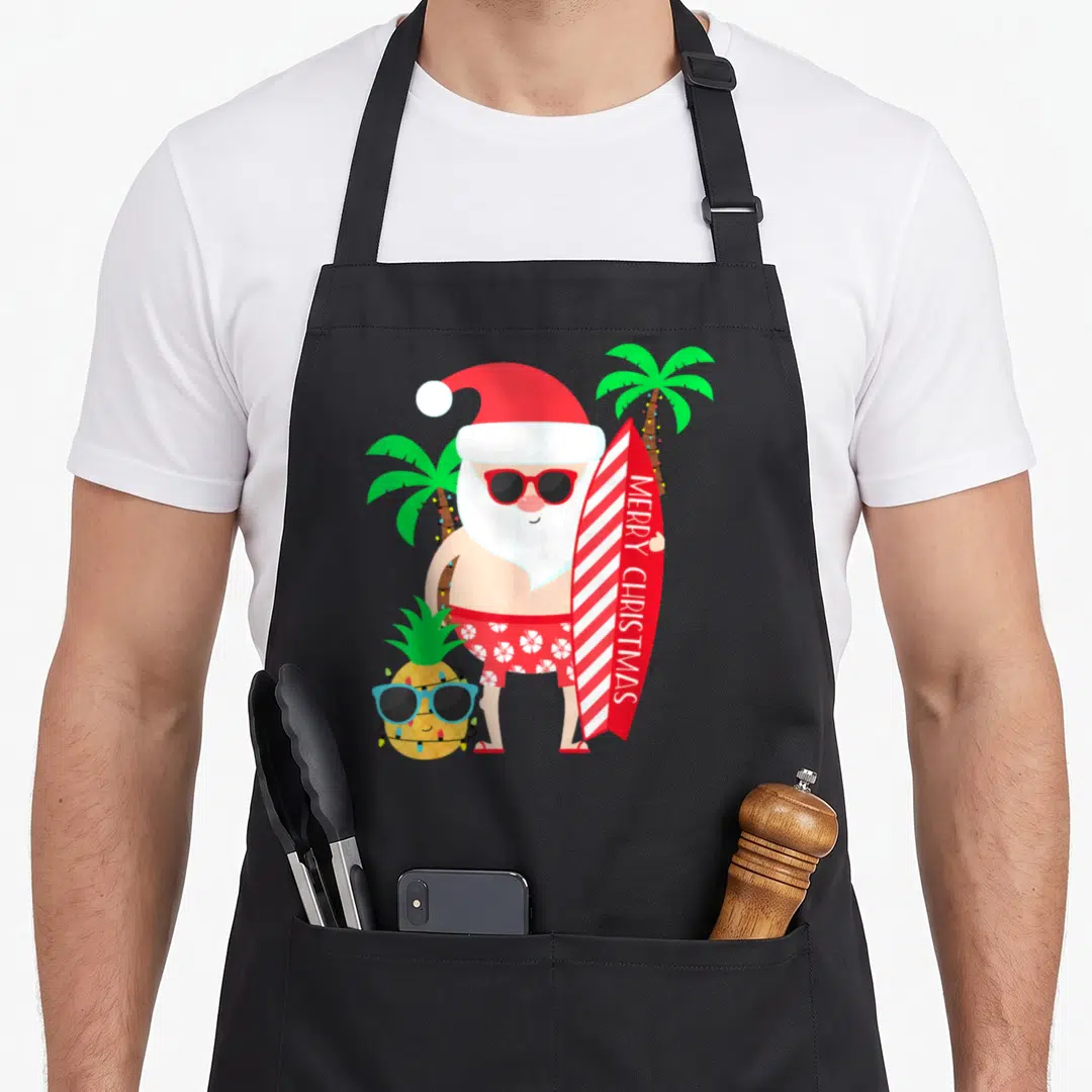 Santa tropical print aprons - Cartoon Santa holding Merry Christmas surfboard next to sunglass-wearing pineapple — shown on a black cotton canvas apron against a white background
