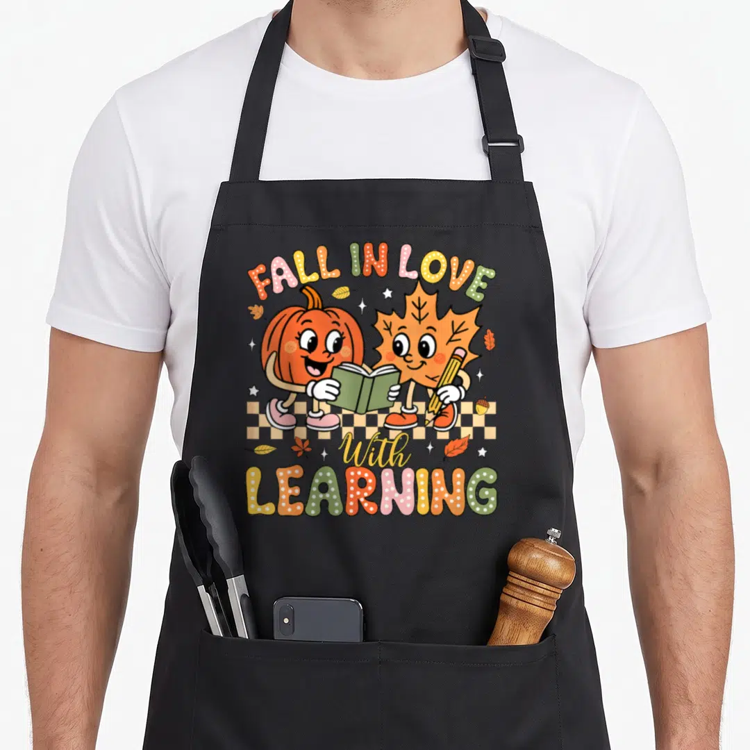Fall teacher apron - Cartoon pumpkin and maple leaf holding book and pencil under arched text on checkered floor — shown on a black cotton canvas apron against a white background