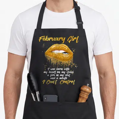 Queen february birthday apron - Gold glittery lips with February Girl text and a multi-line quote about having a fire — shown on a black cotton canvas apron against a white background