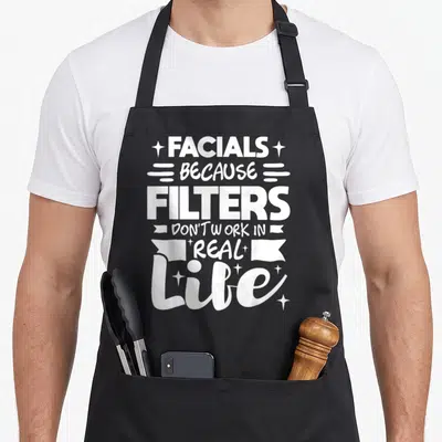 Facial esthetician apron - White typography reading facials because filters don't work in real life with stars — shown on a black cotton canvas apron against a white background