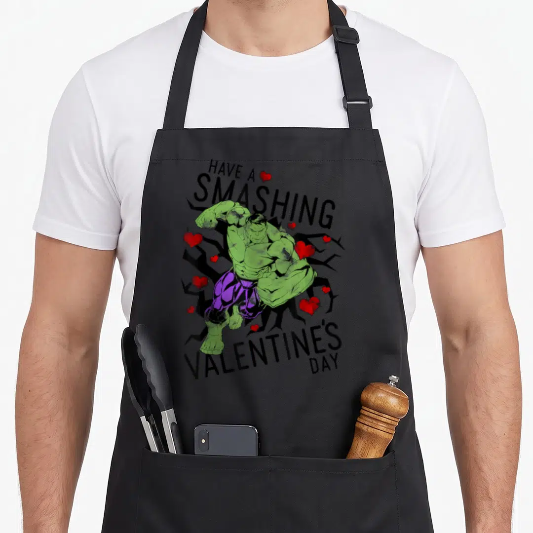 Smashing valentine's day apron - Green character in purple pants punching black cracked background with red hearts — shown on a black cotton canvas apron against a white background