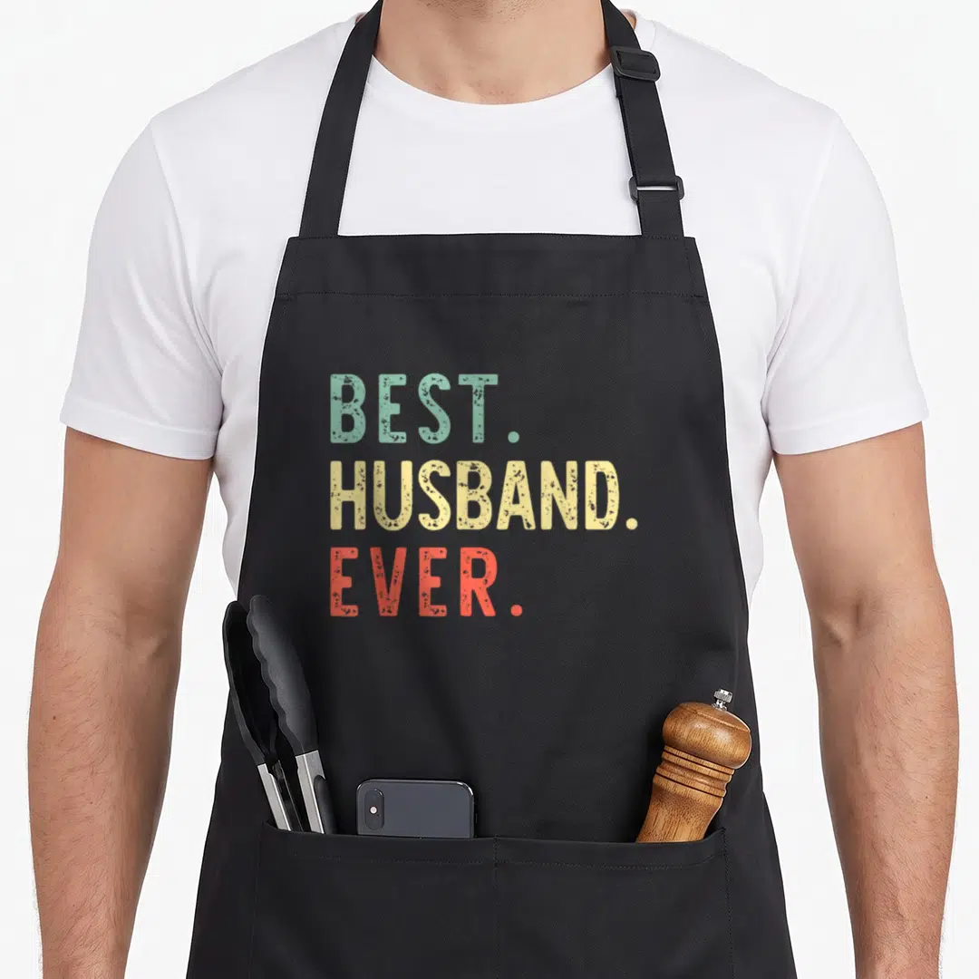 Cool husband apron - Stacked BEST HUSBAND EVER text in green yellow and red distressed typography — shown on a black cotton canvas apron against a white background