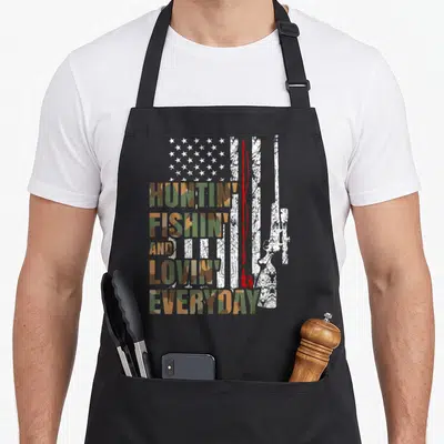 Loving grill hunter apron - Camo text reading Huntin Fishin Lovin Everyday over distressed flag with rifle and rod — shown on a black cotton canvas apron against a white background