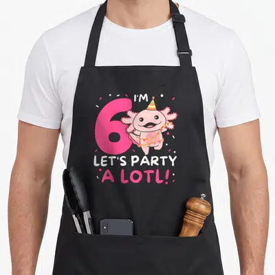 Lotl birthday apron - Pink 6 with cartoon axolotl wearing party hat and bunting surrounded by confetti — shown on a black cotton canvas apron against a white background